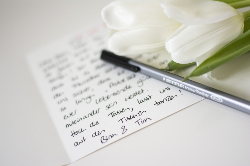 Eine hangeschriebene Karte liegt auf einem Tisch. Auf der Karte liegt ein schwarzer Filzstift der Marke Staedtlder und eine weiße Tulpe.