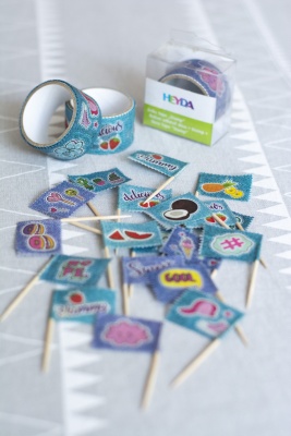 Auf einer weißen Tischdecke, die weiße Wimpelketten zeigt, liegen einige Fähnchen aus Zahnstochern und Washitape. Im Hintergrund sieht man drei Washitaperollen von Heyda.
