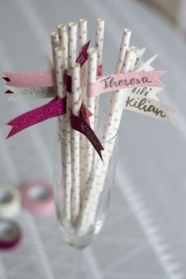 Auf einem Tisch mit weißer Tischdecke steht ein Glas mit gepunkteten Papier-Strohhalmen. Diese wurden mit Fähnchen aus Washitape umklebt und mit Namen beschriftet. Im Hintergrund ist unscharf Washitape in rosa, lila und weiß zu sehen.