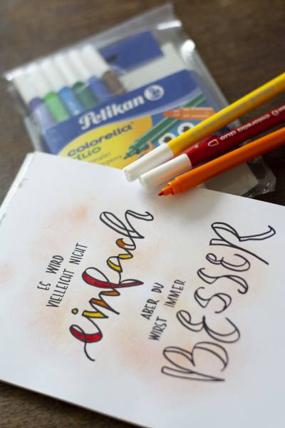 Auf einem Tisch liegt ein Blatt, auf dem der Spruch "Es wird vielleicht nicht einfach, aber du wirst immer besser" gelettert ist. Daneben liegt eine Packung und einzelne Filzstifte der Marke Pelikan