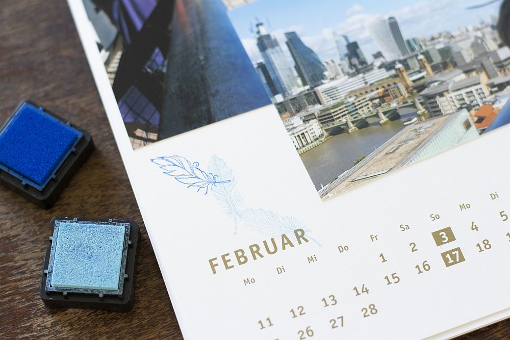 Close-up eines selbstgestalteten Fotokalenders von Heye, der mit blauen Stempeln mit Federmotiv verziert wurde. 