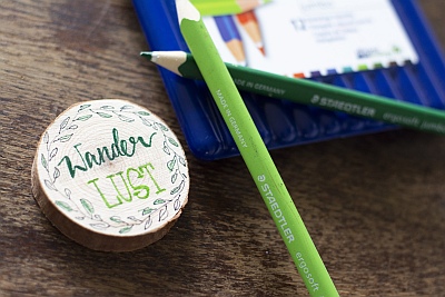 Holzscheibe mit „Wanderlust“-Schriftzug und Laubrahmen, grüne Stifte dabei