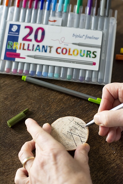 Hand schnitzt Baumzeichnung auf Holzscheibe mit Fineliner und Skalpell, daneben Verpackung von Staedtler triplus Finelinern