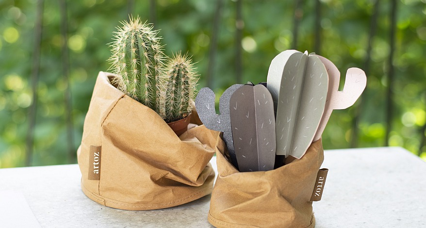 Gebastelte Papierkaktee und echte Kaktee in Braunpapier-Töpfen auf weißem Tisch