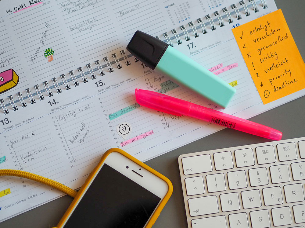 auf einem grauen Untergrund liegt ein aufgeschlagener Tischkalender, auf dem verschiedene Termine eingetragen und markiert sind. Darauf liegen ein türkiser und ein rosa Textmarker. Vor dem Kalender liegen ein Smartphone und eine weiße Tastatur. Auf der Tischkalender der Marke Athesia ist ein Legende mit Erklärungen zu verschiedenen Icons auf ein Post-it notiert.