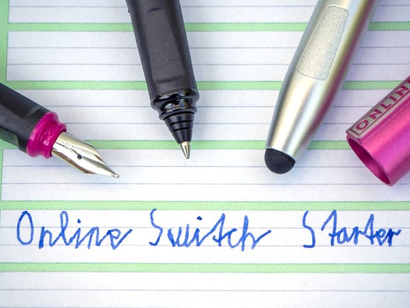 Auf einem Schreiblernheft liegen verschiedene Schreibgeräte von Online. Auf dem Blatt steht "Online Switch Starter" in Schreibschrift, geschrieben mit Tinte.
