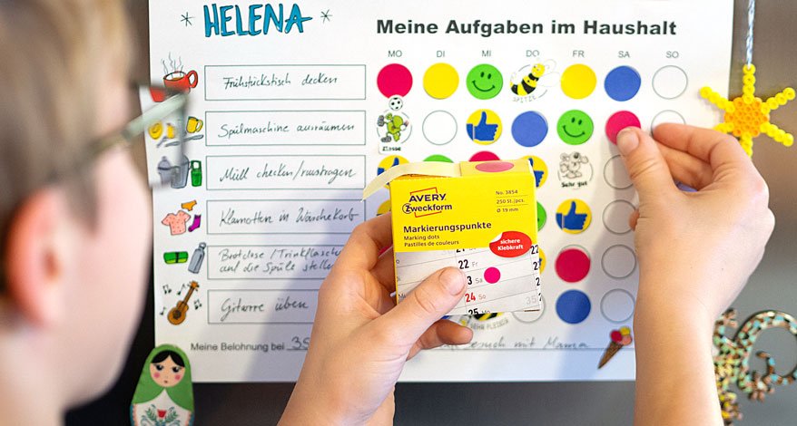 Haushaltsplan für Kinder. Ein Junge klebt einen Markierungspunkt der Marke Avery Zweckform auf die Aufgabentafel.