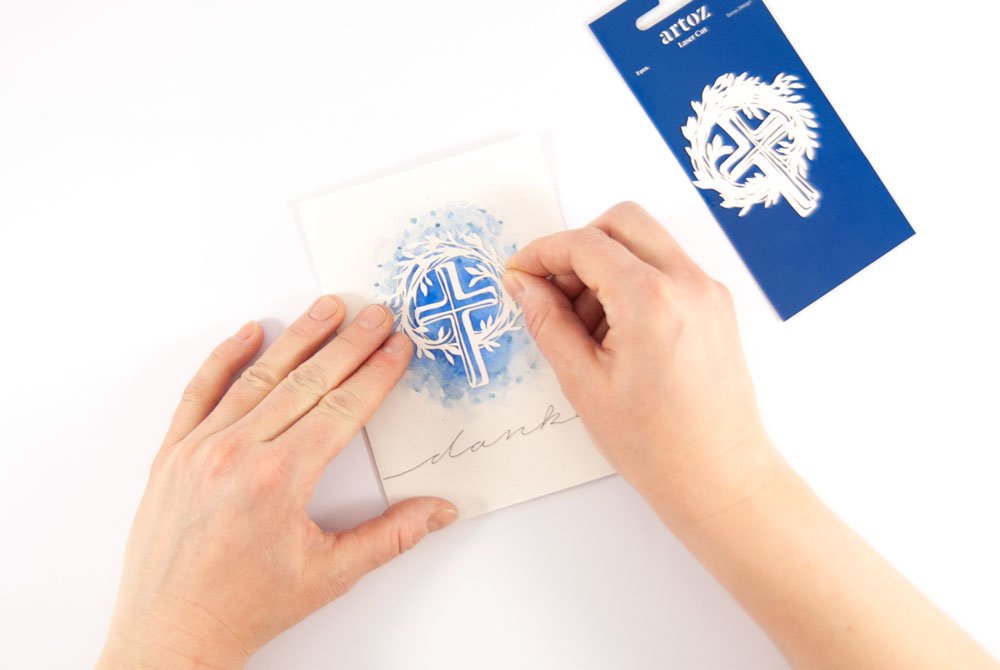 Hand klebt ein weißes Kreuzmotiv blauen Hintergrund einer selbst gestaltete Dankeskarte.