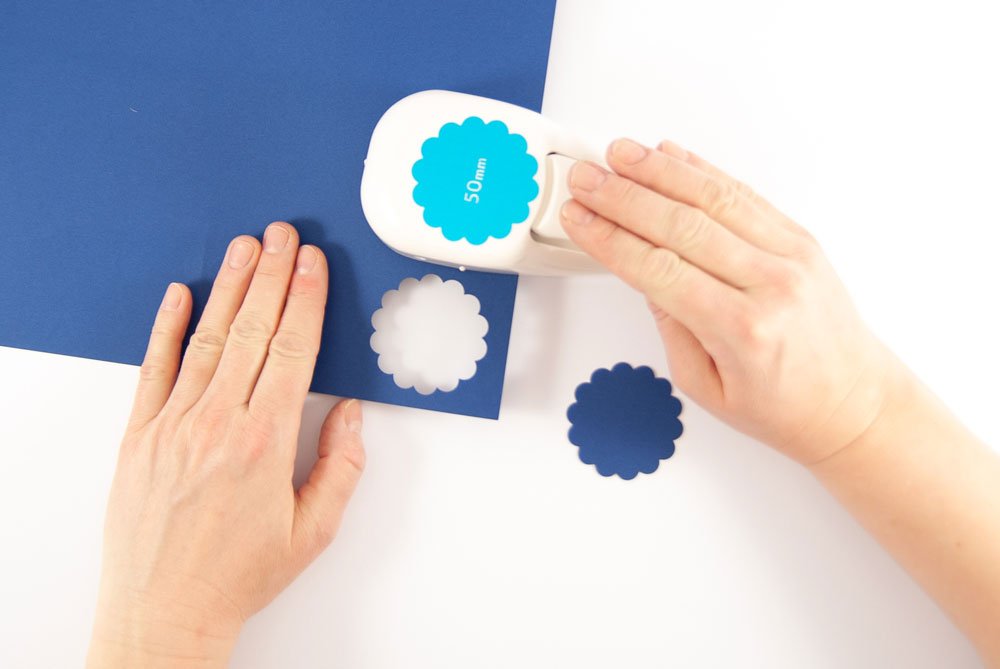 Hand stanzt mit Motivstanzer blaue Kreise aus Papier für eine Dankeskarte.
