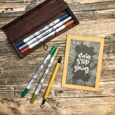 Eine hangeletterte Karte liegt zusammen mit Brushpens und Etui auf einem Holztisch.