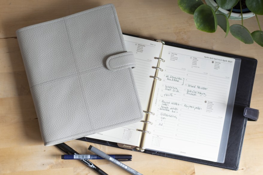 Ein aufgeschlagener sowie ein zugeklappter Planer der Marke Filofax liegen auf einem Tisch. Daneben liegen 3 Stifte.