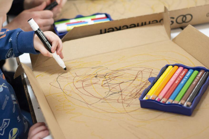 Eine Kinderhand malt mit Stiften aus der Kreativfabrik von Pelikan auf ein Stück Pappe.