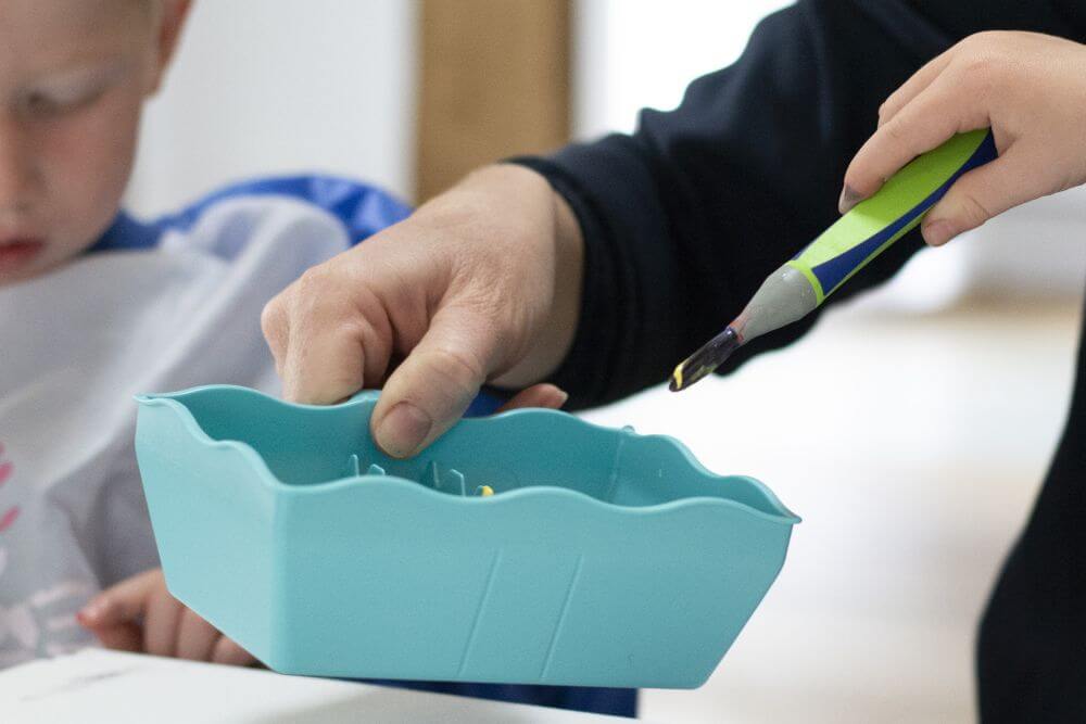 Ein Erwachsener hält einen Wasserbecher aus der Kreativfabrik von Pelikan, eine Kinderhand tunkt einen Pinsel ein.