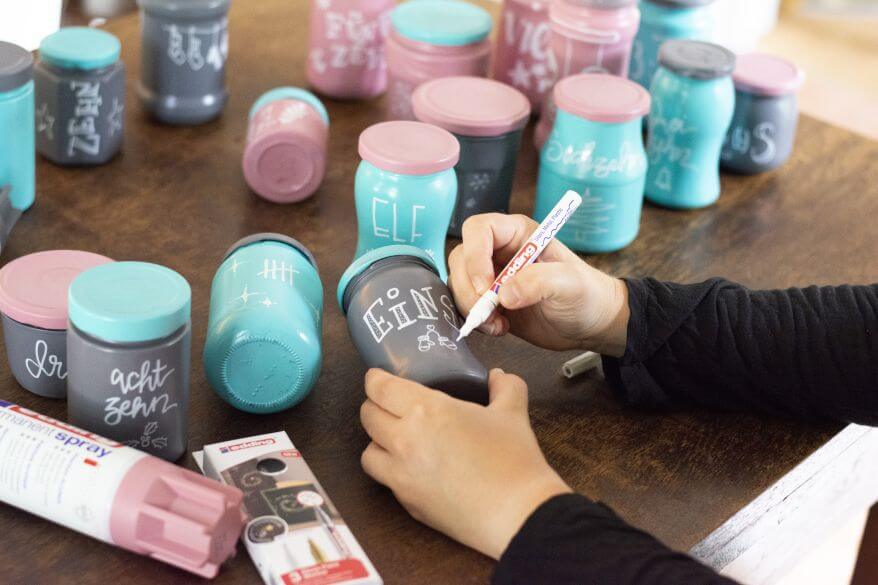 Blick auf einen Tisch, an dem zwei Hände einen Upcycling-Adventskalender aus leeren Marmeladen- und Einmachgläsern gestalten. Die Gläser sind mit Sprühfarbe von Edding in grau, rosa und türkis eingefärbt, die Hände zeichnen mit einem Glanzlackmarker Ziffern und weihnachtliche Zeichnungen auf.