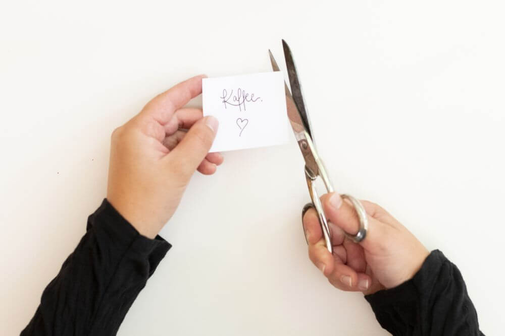 Auf einen weißen Zettel ist das Wort "Kaffee" gelettert. Darunter ist ein Herz gezeichnet. Der Zettel wird von einer Hand gehalten und mit der andern Hand durch eine Schere zurechtgeschnitten.Der Hintergrund ist weiß.
