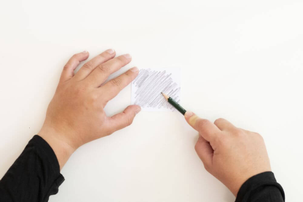 Ein weißer Zettel liegt auf einem weißen Untergrund. Er wird durch eine Hand festgehalten. Mit der anderen Hand wird mit Hilfe eines Bleistiftes das Papier schraffiert.