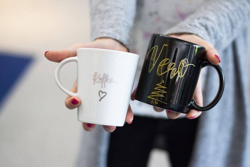 Eine Frau mit einem grauen Oberteil hält zwei Tassen in die Kamera. Diese wurden mit Metallic-Pens von Faber Castell belettert. Auf der einen steht "Kaffee" mit einem Herz darunter. Die andere Tasse ist schwarz und darauf ist der Name "Vero" gelettert.Darunter wurde ein minimalistischer Tannenbaum gemalt.