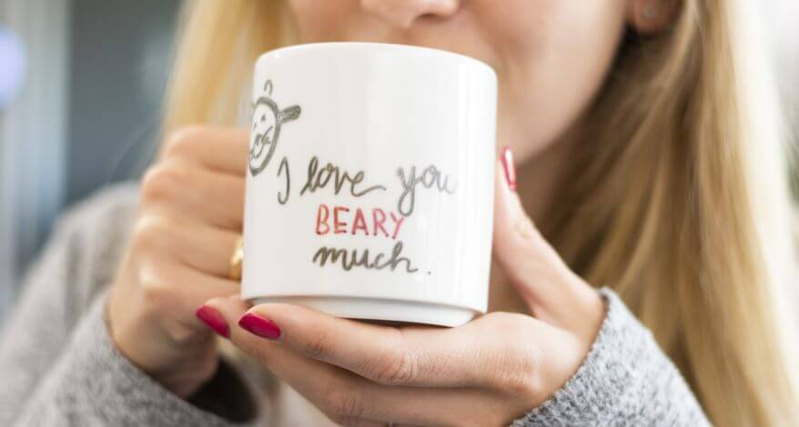 Eine Frau mit blonden Haaren und einem grauen Pullover hält sich eine Tasse an den Mund. Diese wurde mit den Worten "I love you beary much." belettert. Außerdem ist eine kleine Zeichnung eines Bären zu erkenne.