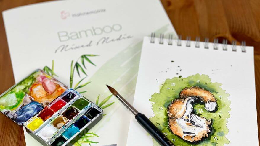 Bamboo-Mix-Media-Papier aus der Serie Natural Line von Hahnemühle liegt als Malblock auf einem Tisch. Im Vordergrund sieht man einen Kasten mit Aquarellfarben und einer verspielten Aquarellillustration mit Fuchs. Foto und Illustration von Verena Knabe.