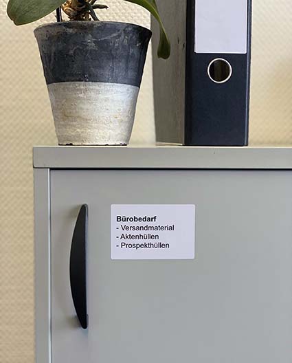 Auf einen Schrank stehen eine Blume in einem schwarz-weißen Topf sowie ein schwarzer Ordner. Der Schrank im mit einem Etikett der Marke Avery Zweckform mit den Worten Bürobedarf: Hüllen und Versandmaterial beschriftet.