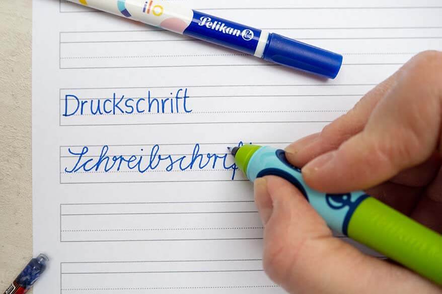 Eine Hand schreibt mit einem griffix Schreiblern-Tintenroller von Pelikan die Worte Druckschrift in Druckschrift und Schreibschrift in Schreibschrift auf ein Schreiblernblatt. Im Bild sieht man außerdem einen Tintenlöscher und eine Tintenpatrone. Foto: Johanna Rundel 