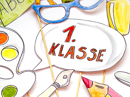 Verschiedene Fotoprops für Einschulungsfotos liegen auf einem weißen Untergrund. Zu sehen sind: eine Sprechblase, in der "1. Klasse" steht, eine blaue Brille, eine bunter Wasserfarbkasten, ein Buntstift und ein gelber Textmarker. Der für die Props verwendete Fotokarton ist von der Marke Heyda, die Holzstäbe sind von Knorr-Prandell. Foto: Johanna Rundel