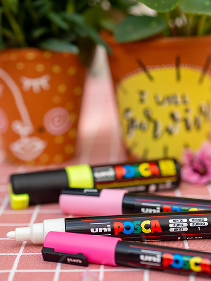 Auf einer rosa-weiß karierten Tischdecke liegen vier Marker der Marke POSCA. Im Hintergrund sind zwei Blumentöpfe aus Ton zu sehen, die mit diesen bunten Markern angemalt wurden. Foto: Johanna Rundel