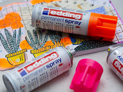 Auf einem bunten Art Journal Kunstwerk in Mixed-Media Technik liegen drei Acrylsprays der Marke Edding. Die Sprühdosen enthalten folgende Farben: Neonpink, Neongelb und Neongrün. Der Untergrund ist hell. Foto: Johanna Rundel