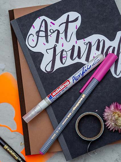 Ein braunes und ein schwarzes Notizbuch liegen auf einem hellen Untergrund. Auf dem schwarzen Notizbuch ist Art Journal gelettert. Auf dem Stapel liegen zwei Stifte der Marke Edding sowie eine Rolle Washitape und eine Trockenblume. Der Untergrund ist hell. Foto: Johanna Rundel