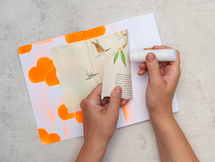 Jemand klebt mit einem Klebestift einen Ausschnitt aus einem Magazin auf die Seiten eines Art Journals im Mixed-Media-Stil. Darauf sind fliegende Gänse und Wolken zu sehen. Foto: Johanna Rundel