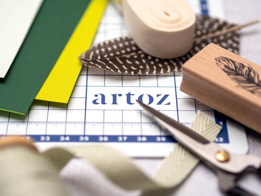 Auf einer Schneidematte von Artoz liegen verschiedene Bastelmaterialien, wie Papier in Grüntönen, Federn, eine Schere, helles Baumwollband und ein Stempel mit Federmotiv. Foto: Johanna Rundel