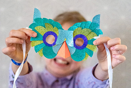 Ein Mädchen im Grundschulalter hält eine selbstgebastelte Vogelmaske in die Kamera. Diese ist mit bunten Glitzerpapieren in Grüntönen der Marke Artoz beklebt. Foto: Johanna Rundel