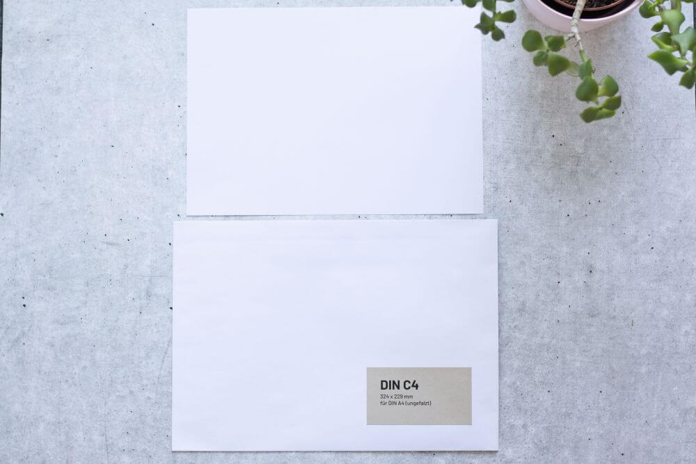 Auf grauem Grund liegt ein DIN A4-Papier und eine dazu passende Versandtasche DIN C4. Auf der Versandtasche ist ein Aufkleber aufgebracht mit folgenden Infos: DIN C4, 324x229 Millimeter, für DIN A4 (ungefalzt). Rechts oben ragt der Teil einer Pflanze ins Bild.