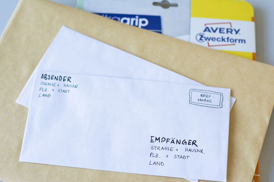 Mehrere Briefumschläge und Versandtaschen liegen aufeinander gestapelt. Ganz oben liegt ein Briefumschlag im Format DIN lang, auf dem in schwarzer Druckschrift geschrieben steht, wohin welches Element auf dem Briefumschlag kommt: Oben links Absender mit Straße, Hausnummer, Postleitzahl, Stadt und Land. Unten rechts Empfänger mit Straße, Hausnummer, Postleitzahl, Stadt und Land. Oben rechts ist an der Stelle, wo die Briefmarke hinkommt, ein Icon dafür eingezeichnet. Im Hintergrund sieht man die Verpackung von Adressetiketten von Avery Zweckform.