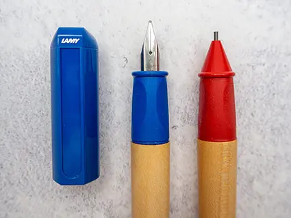 Ein blauer Schreiblernfüller und ein roter Schreiblernbleistift von Lamy liegen nebeneinander. Links daneben liegt der blaue Füllerdeckel.
