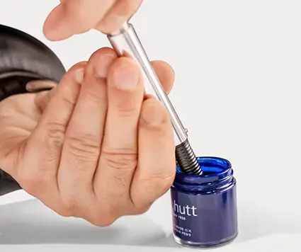 Jemand zieht blaue Tinte aus einem Tintenfässchen in einen silbernen Füller von Otto Hutt. Der Hintergrund ist weiß.