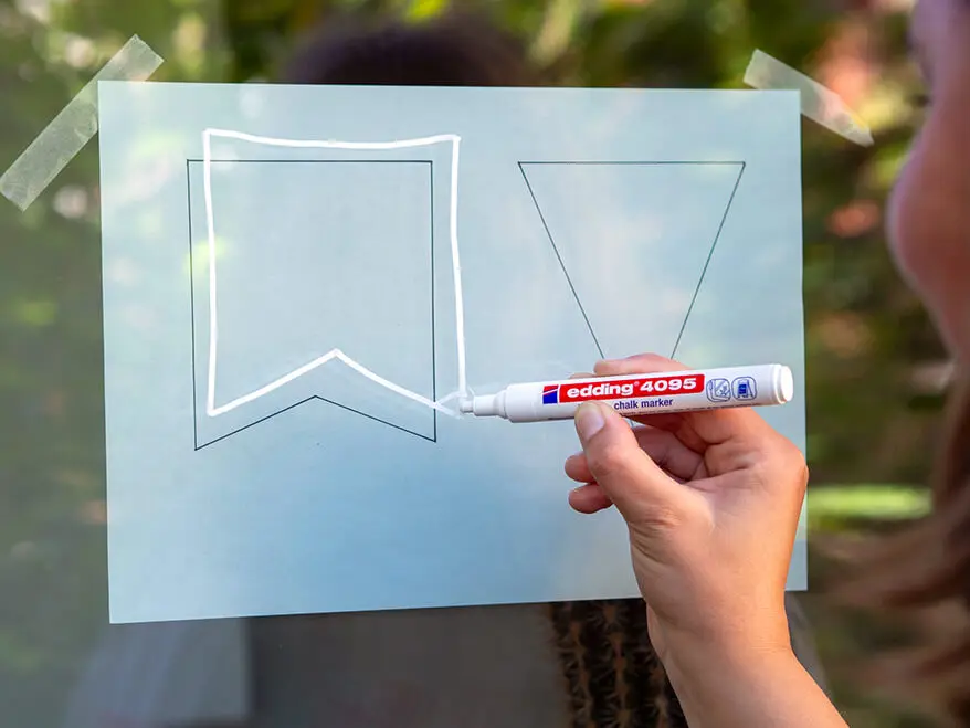 Jemand malt mit einem weißen Kreidemarker von edding eine Wimpel-Vorlage von einer Schablone auf eine Fensterscheibe. Foto: Johanna Rundel