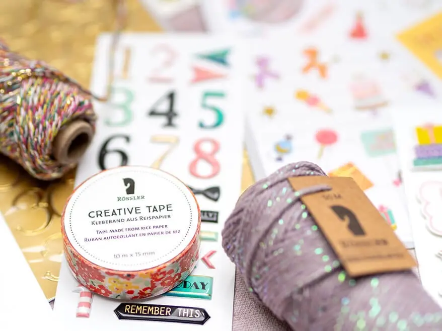 Viele bunte Bastelprodukte, wie Aufkleber, Washitape und Bäckergarn von Rössler, liegen verstreut auf einer rosa Arbeitsfläche. Foto: Johanna Rundel 