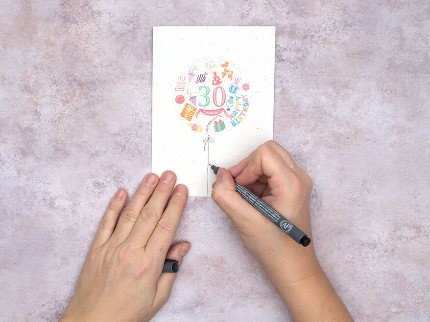 Jemand zieht mit einem schwarzen Fineliner eine dünne Linie auf einer gestalteten Blanko-Klappkarte von Rössler. Foto: Johanna Rundel 