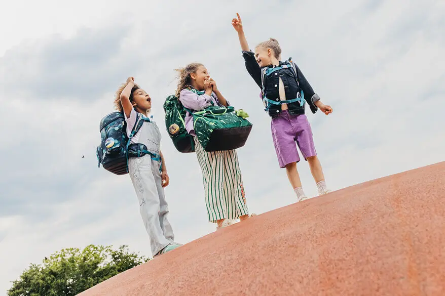 Drei Mädchen im Grundschulalter stehen mit Ihren Schulranzen und Sporttaschen von ergobag auf einer Anhöhe und blicken in den Himmel.