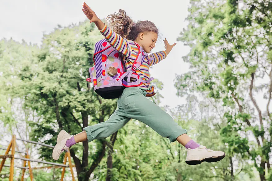 Ein Mädchen im Grundschulalter springt mit ihrem Schulranzen von ergobag auf dem Rücken beschwingt über einen Spielplatz im Grünen.