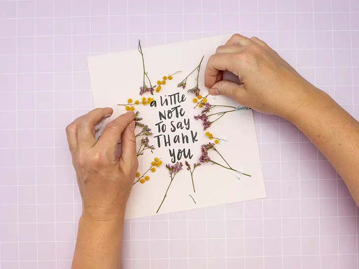 Jemand legt Trockenblumen um ein, mit einem Kreativmarker von Faber-Castell geschriebenes, Lettering auf einem rosa Fotokarton. Foto: Johanna Rundel