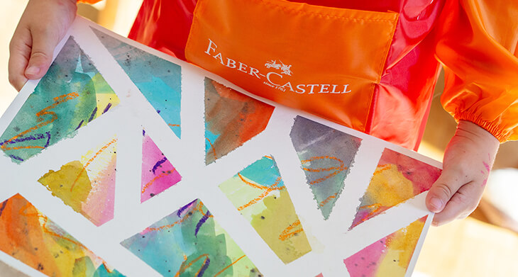  Ein Kleinkind in einer orange-roten Malerschürze von Faber-Castell hält ein Tape-Resist-Art-Kunstwerk in die Kamera. Foto: Johanna Rundel