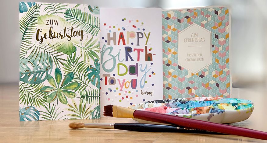 3 bunte Geburtstagskarten aufgestellt auf einem Tisch, vor den Karten 2 Pinsel und eine Farbpalette