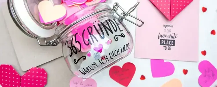 Ein belettertes Einmachglas auf einem Tisch, drumherum liegen rosa und rote Papierherzen und eine gebastelte Karte. 