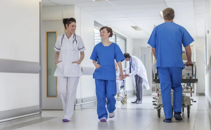 Das Bild zeigt einen Krankenhausflur. Eine Ärztin und eine Krankenschwester unterhalten sich. Im Hintergrund wird ein Patient auf einer Bahre von einer anderen medizinischen Fachkraft betreut.