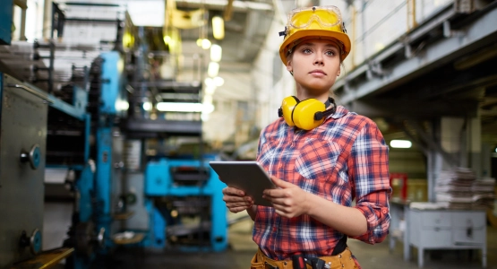 Junge Frau mit Schutzhelm und Gehörschutz hält Tablet in einer Fabrikumgebung.