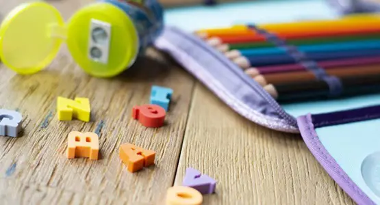 Auf einem Holztisch liegt ein aufgeklapptes Federmäppchen, ein Spitzer und bunte Radiergummis in Buchstabenform.