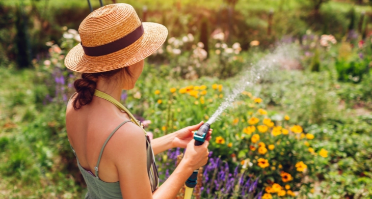 Frau mit Strohhut gießt bunte Blumen im Garten mit einem Gartenschlauch. 