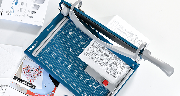 "Das Bild zeigt ein Dahle Hebelschneider in Blau auf einem Tisch mit. Das Gerät hat eine große Schneideklinge, einen ergonomischen Griff und eine Bedruckung auf der Tischfläche, die es ermöglicht, präzise Formate zu schneiden. Mehrere Blätter Papier liegen auf dem Schneidetisch und unter dem Hebelarm des Schneiders.“ 